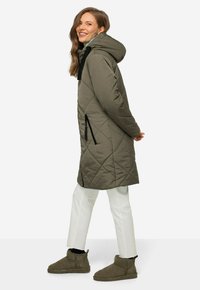 Cappotto lungo in khaki trapuntato con cappuccio, tasche frontali con zip e cordini regolabili. Abbinato a pantaloni bianchi e stivali alla caviglia color oliva.