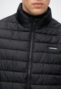 Άνδρας που φοράει μαύρο καπιτονέ μπουφάν Jack & Jones με φερμουάρ και όρθιο γιακά, κομμένο στα χείλη και τους ώμους.