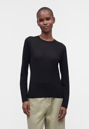 Person mit kurzen Haaren, die ein eng anliegendes schwarzes Langarmshirt und lockere beigefarbene Hosen trägt, steht vor einem schlichten hellen Hintergrund.
