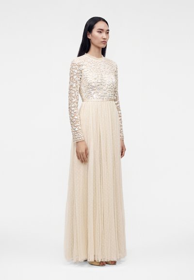 Femme aux longs cheveux foncés portant une robe beige à manches longues, longue jusqu'au sol, avec des motifs floraux et à pois texturés, debout devant un fond uni.