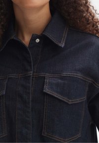 Mörk denimskjorta med spetsig krage, två framfickor och kontrasterande orange sömmar. Har silverfärgade tryckknappar. Standard passform.