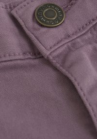 Ljust lila tyg med en synlig sydd söm, prydd med en rund metallknapp med präglad text och ett stjärnmönster.