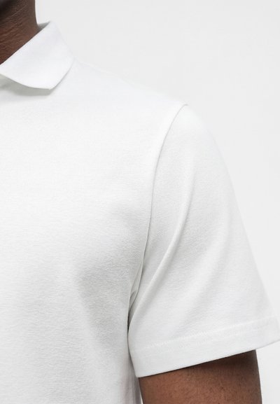 Gros plan d'une personne portant un polo blanc uni à manches courtes montrant les détails du col et des manches.