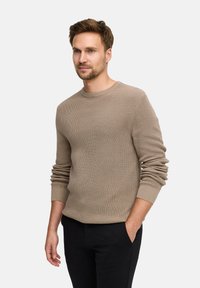 Beige, strukturiertes Strickpullover mit Rundhalsausschnitt und langen Ärmeln, kombiniert mit schwarzen Hosen. Der Pullover hat gerippte Bündchen und Saum.