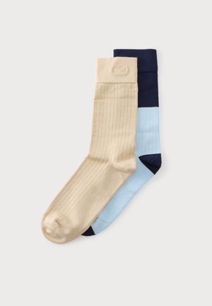 Une chaussette beige côtelée chevauche une chaussette à panneaux bleu et marine sur un fond gris clair uni.