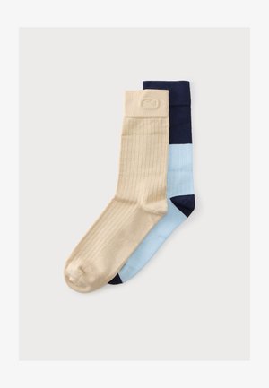 Une chaussette beige côtelée chevauche une chaussette à panneaux bleu et marine sur un fond gris clair uni.