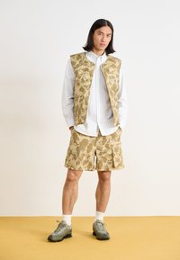 Lättviktsväst och shorts i kamouflagemönster i beige toner. Vitrandig skjorta under. Olivgröna sneakers med texturerad sula.
