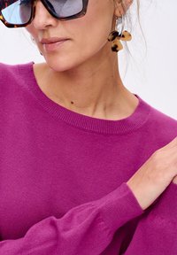 Kobieta w dużych okularach przeciwsłonecznych w stylu żółwim, z kolczykami w kształcie kółek z akcentami w postaci kokardek w stylu żółwim oraz jasnofioletowym swetrem z dzianiny.