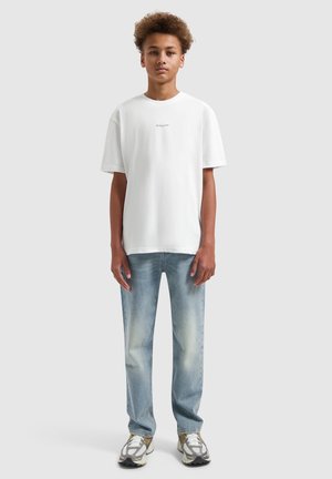 Tiener draagt wit T-shirt met korte mouwen, lichtblauwe vervaagde spijkerbroek en grijze sneakers, staand tegen een effen lichte achtergrond.