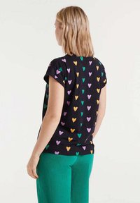 Camiseta negra de manga corta, con patrones de corazones coloridos en púrpura, verde y naranja, combinada con pantalones verdes texturizados.