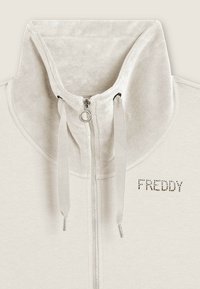 Felpa leggera beige con zip e colletto alto, caratterizzata da una texture morbida, cordini e la scritta "FREDDY" in strass sulla parte anteriore.