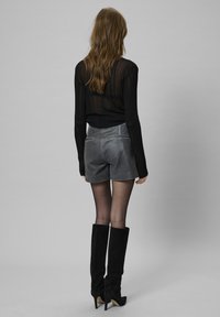 Črna prosojna majica z dolgimi rokavi, sivi semiš kratki hlače z zadnjimi žepi in črne hlačne škornje do kolen z ostro peto na sivi ozadju.