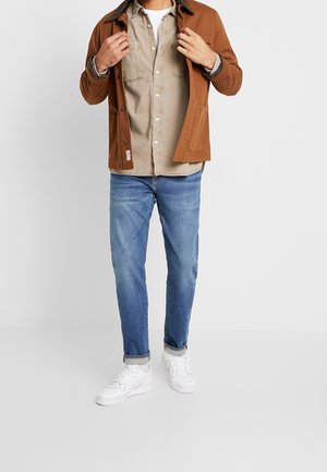 Brun bomullsjacka över en beige skjorta med knappar, blå jeans i denim och vita sneakers; avslappnad lager-på-lager-stil med strukturerade designelement.