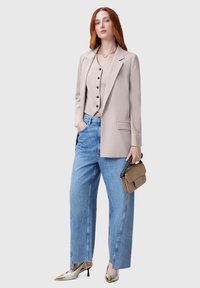 Beige bleiseri yang disesuaikan di atas atasan ringan dengan kancing, dipadukan dengan celana jeans biru muda yang longgar dan sepatu hak runcing emas, sambil memegang tas tangan kecil berwarna cokelat muda.