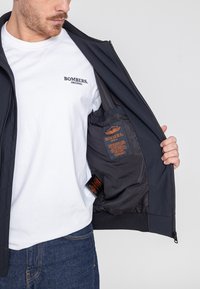 Marineblauer Bomberjacke mit einem gesteppten Design, kontrastierendem Innenfutter und orangem Etikett. Getragen über einem weißen T-Shirt und blauen Jeans.