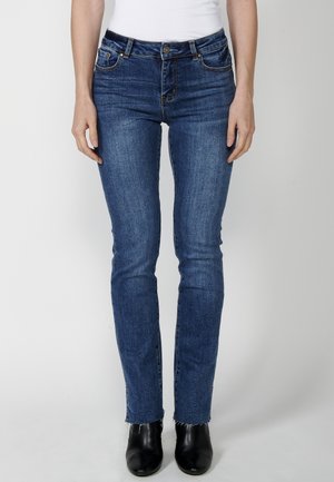 Personne portant un jean slim bleu en denim avec des ourlets effilochés, des bottines noires et un haut blanc rentré, debout sur un fond uni.