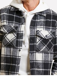 Veste-chemise à carreaux pour hommes en noir et blanc, fabriquée dans un tissu doux. Comprend deux poches poitrine et une fermeture boutonnée sur le devant.