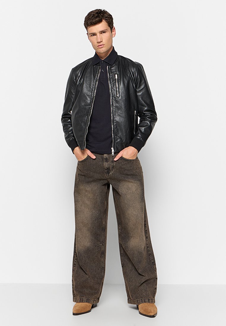 Zwarte leren bomberjack met een ritssluiting en zilveren accenten, gecombineerd met ruime bruine denim broeken en tannenkleurige suède schoenen.