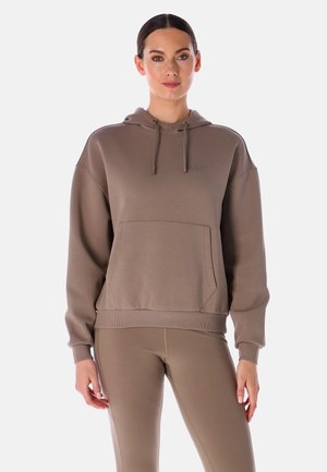 Une femme porte un sweat à capuche taupe avec une poche sur le devant et un legging assorti, sur un fond blanc uni.