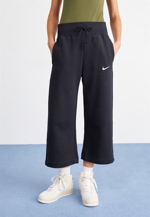 Person, der schwarze weite, gekürzte Sweatpants mit Nike-Logo, weiße High-Top-Sneaker und ein olivgrünes Oberteil trägt und auf einem blauen Boden steht.