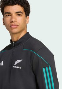 Crna zip jakna s tirkiznim detaljima, s logotipom All Blacks i bijelim Adidas brandom. Izrađena od glatkog materijala s visokim ovratnikom.
