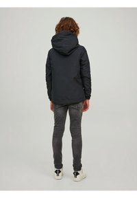 Dziecko stojące tyłem do kamery, ubranie w czarną kurtkę z kapturem, wyblakłe szare dżinsy i białe trampki, na jednolitym szarym tle.