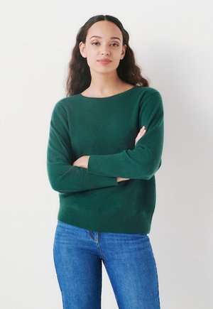 Pull en tricot vert avec un col large et des manches longues, associé à un jean bleu. Le tissu semble doux avec une texture subtile.
