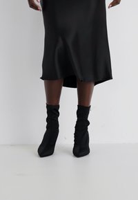 Vestido de satén negro con una silueta fluida, dobladillo a la altura de la rodilla y un elegante drapeado, combinado con botas de tobillo negras y holgadas.