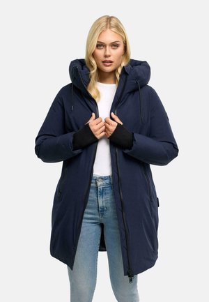 Donna con capelli biondi che indossa un cappotto invernale blu navy con cappuccio, camicia bianca e jeans azzurri, tenendo la zip della giacca vicino al petto.