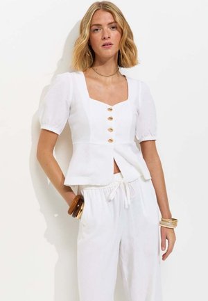 Femme blonde portant un chemisier blanc à manches courtes boutonné et un pantalon blanc à cordon, accessoirisée de bracelets dorés et bruns.