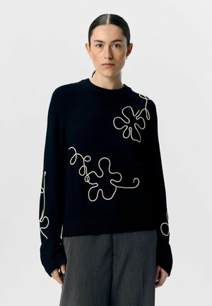 Zwarte gebreide trui met beige abstracte geborduurde ontwerpen, lange mouwen met verlengde manchet en een ronde halslijn.