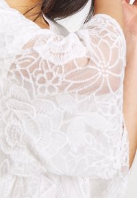 Sciarpa bianca in pizzo con motivi floreali, texture semi-trasparente e caduta morbida, con maniche corte e dettagli di design intricati.