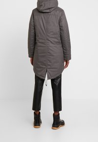 Parka grigia lunga con cappuccio e orlo arrotondato. Presenta una texture liscia e uno spacco posteriore, abbinata a pantaloni in pelle nera e stivali.