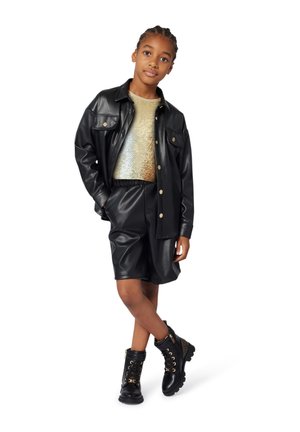 Fille debout avec les jambes croisées, portant une chemise et un short en simili cuir noir, un haut en sequins dorés, et des bottes noires à lacets avec des détails dorés.