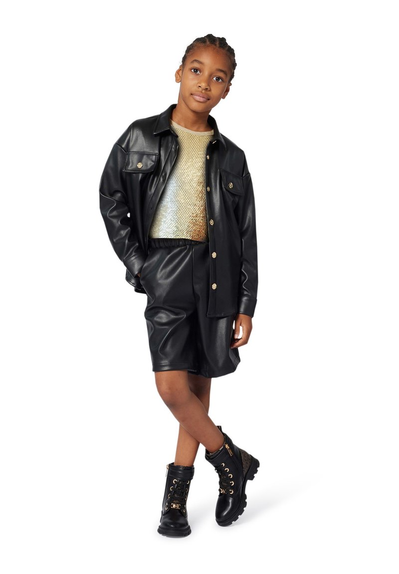 Ragazza in piedi con le gambe incrociate, che indossa una camicia e dei pantaloni corti in finta pelle nera, un top di paillettes dorate e stivali neri stringati con dettagli dorati.