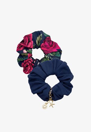 Kobieta o długich włosach, ubrana w granatowy bikini w kwiaty z wiązaniem na szyi i dopasowane majtki, uśmiechająca się z założonymi na piersi ramionami.