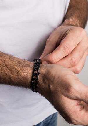 Black chain bracelet made of smooth, glossy material. Features interlocking links, worn on a wrist with light hair.