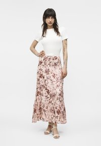 Kvinde med langt mørkt hår iført hvid kortærmet top, lang lyserød nederdel med blomsterprint og hvide sandaler med stropper, stående med hånden på hoften.