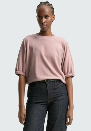 Un haut rose clair à manches courtes avec un col rond et des détails de manches froncées, associé à un jean en denim foncé avec une ceinture.