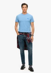 Camiseta de manga corta azul de brezo, jeans oscuros, mocasines negros, camisa de cuadros atada a la cintura, reloj en la muñeca izquierda.