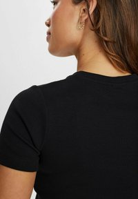 Tricou negru cu maneci scurte, cu un guler rotund. Vedere laterală care arată textura netedă și designul ajustat. Cercei hoop aurii vizibili.