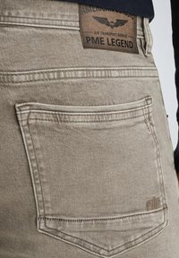 Jeans de mezclilla beige con un bolsillo trasero que presenta un contorno acolchado y una etiqueta de cuero marrón con la inscripción "PME Legend" y un diseño de alas.