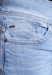 Světle modré denimové džíny mají zadní kapsu s viditelným logem "RAW" a jemnými detaily oděru a šití.