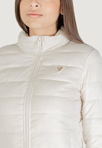 Witte pufferjack met een hoge kraag, horizontaal gequilt patroon, rits aan de voorkant en een klein driehoekig logo detail.