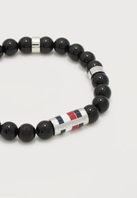 Μαύρο βραχιόλι με χάντρες και ασημένια κυλινδρική χάντρα με ρίγες κόκκινου, λευκού και μπλε, χαραγμένη με την επιγραφή "HILFIGER" σε λευκό φόντο.