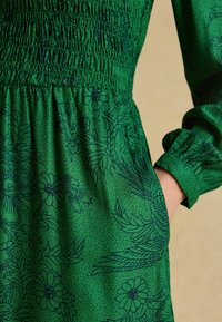 Zelené šaty so smockovaným pásom a dlhými rukávmi, s tmavými kvetinovými vzormi; ruka je položená v vrecku na pravej strane.