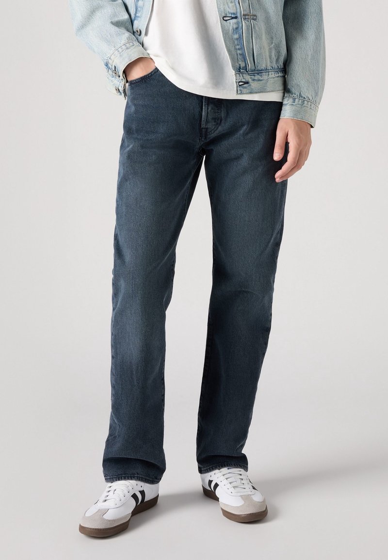 Dunkelblaue Jeans aus Denim mit geradem Schnitt, detaillierten Nähten und einem Fünf-Taschen-Design, kombiniert mit weißen Sneakers und einer hellen Jeansjacke.