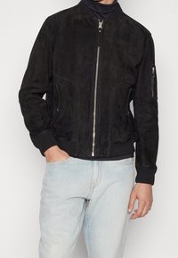 Giacca bomber nera in suede con zip frontale, polsini a costine e tasche laterali. Texture liscia, vestibilità strutturata e design minimalista.