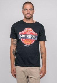 T-shirt en coton noir avec un impression graphique colorée d'une étiquette d'huile moteur vintage, à manches courtes et col rond.