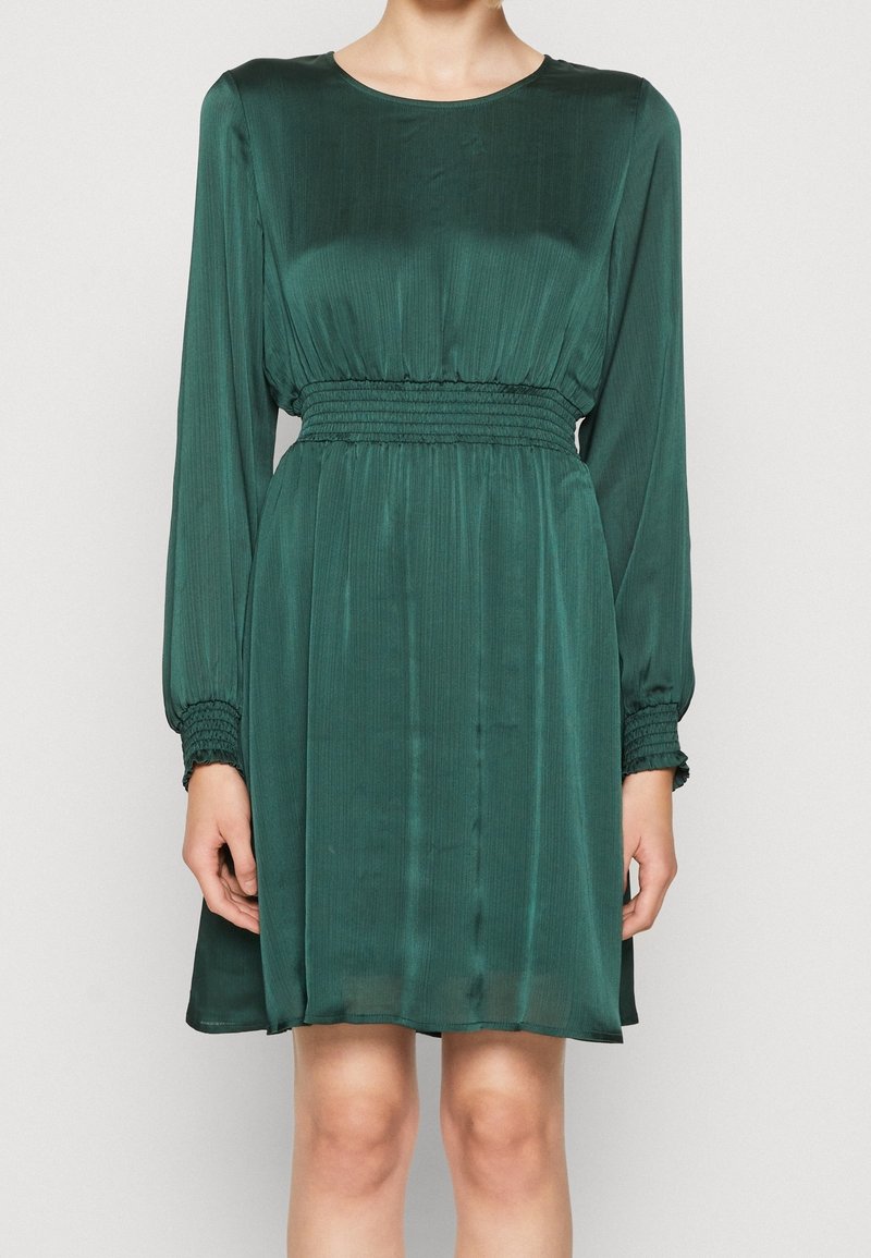 Femme portant une robe verte foncée à manches longues avec une taille cintrée et froncée ainsi que des poignets assortis froncés.